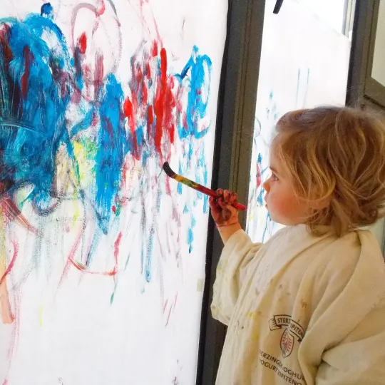 bambina impugna un pennello colorando un grande foglio verticale dipinto con molti colori 