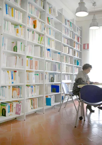 Interno della Biblioteca con libreria a parete ricolma di libri, un'ampia finestra luminosa e tre utenti in consultazione 